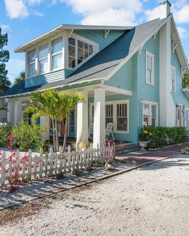 Turquoise and white cottage with pitched roof, white picket fence, palm trees, and American flag in front yard