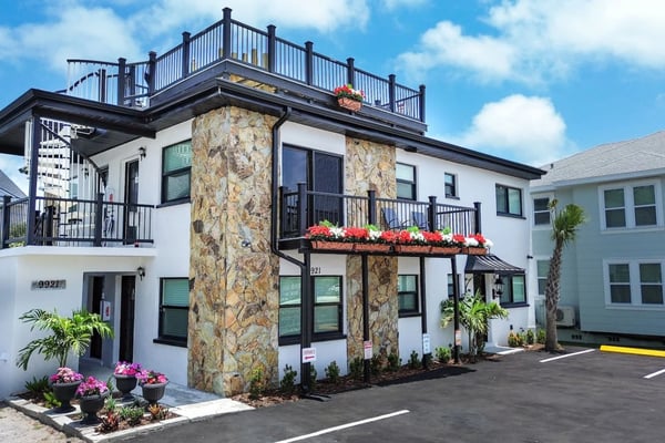 Modern white vacation rental with stone corner tower, rooftop deck, red and pink flowers, and palm trees under blue sky